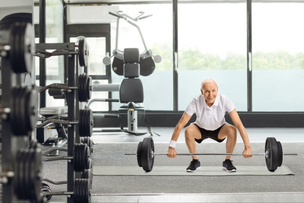 An older adult man bends his hips and knees to lift a barbell in a gym.