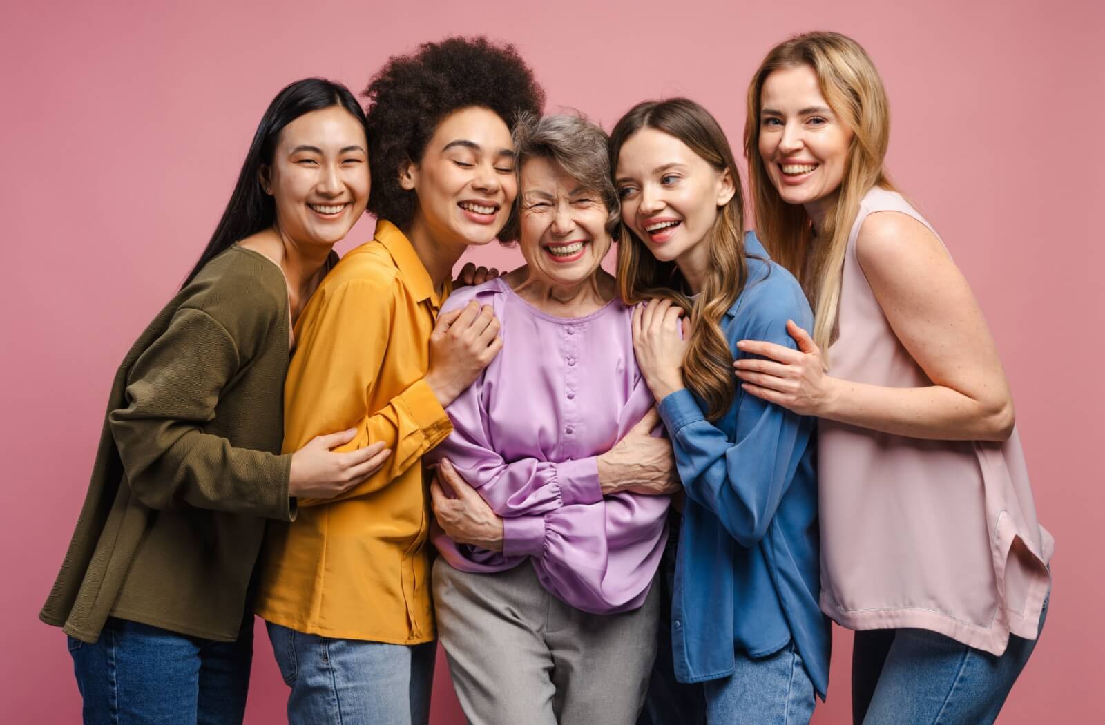 Against a pink background, four young adults crowd around an older adult, representing an intergenerational friendship